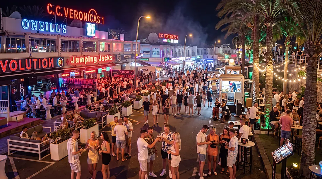 Crowded beachfront promenade at night with neon signs and palm trees
