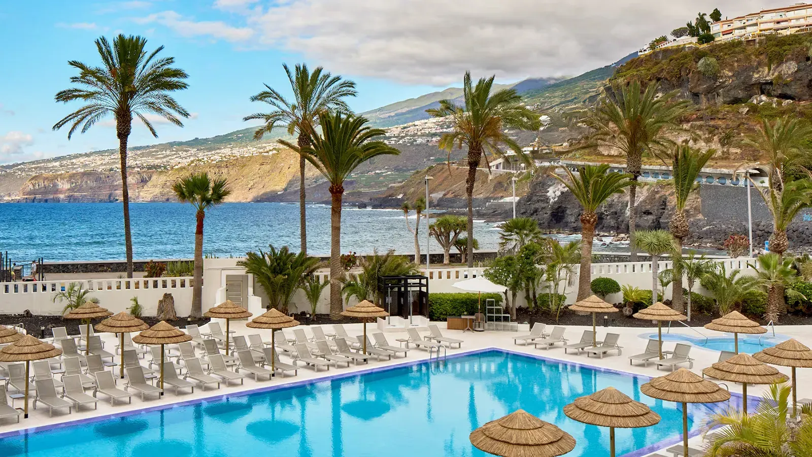 Resort pool with ocean view, palm trees, and volcanic cliffs