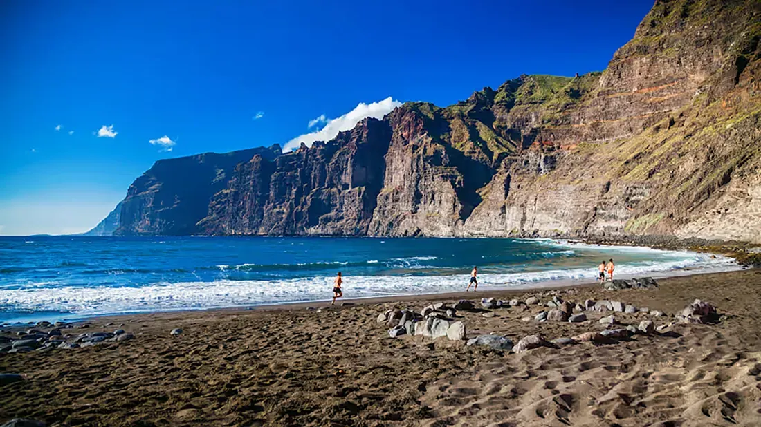 Swimmers and beachgoers on black sand beach beneath towering coastal cliffs