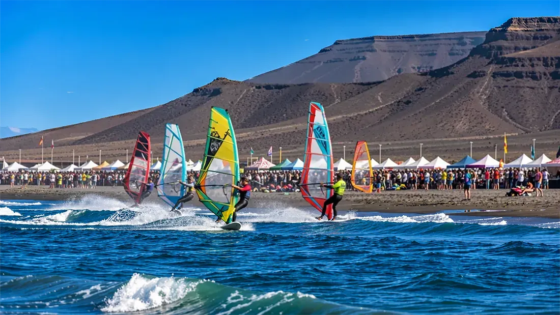 Windsurfers competing in colorful sails near crowded beach festival tents