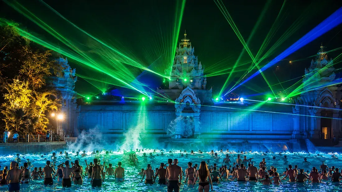 Crowds swimming in illuminated thermal pool with laser light show and ornate buildings