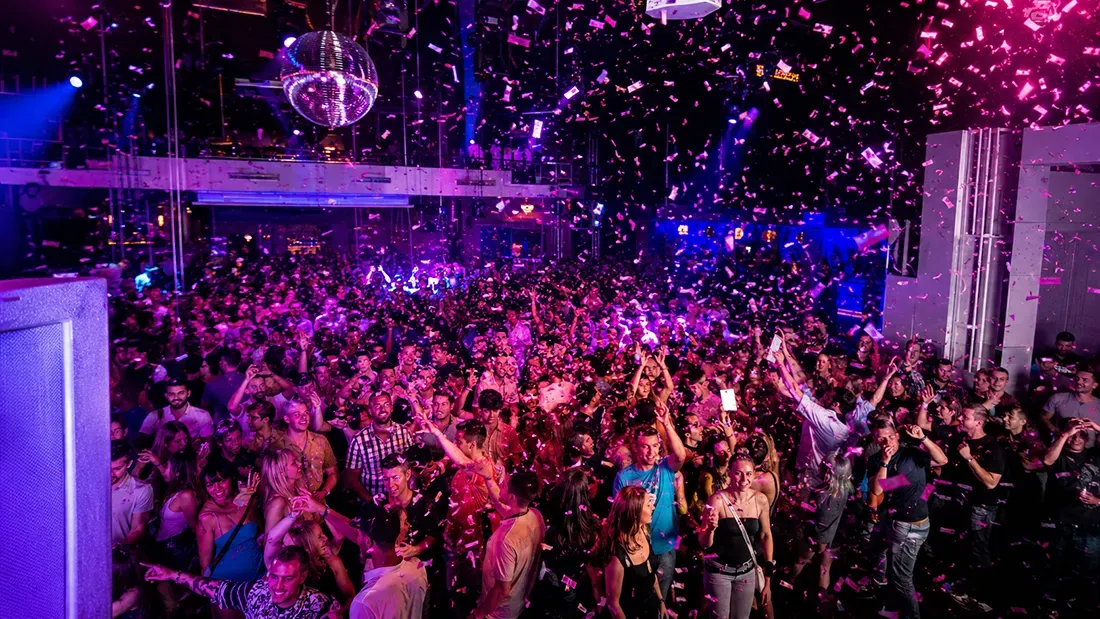 Large crowd dancing under purple lights with confetti falling indoors