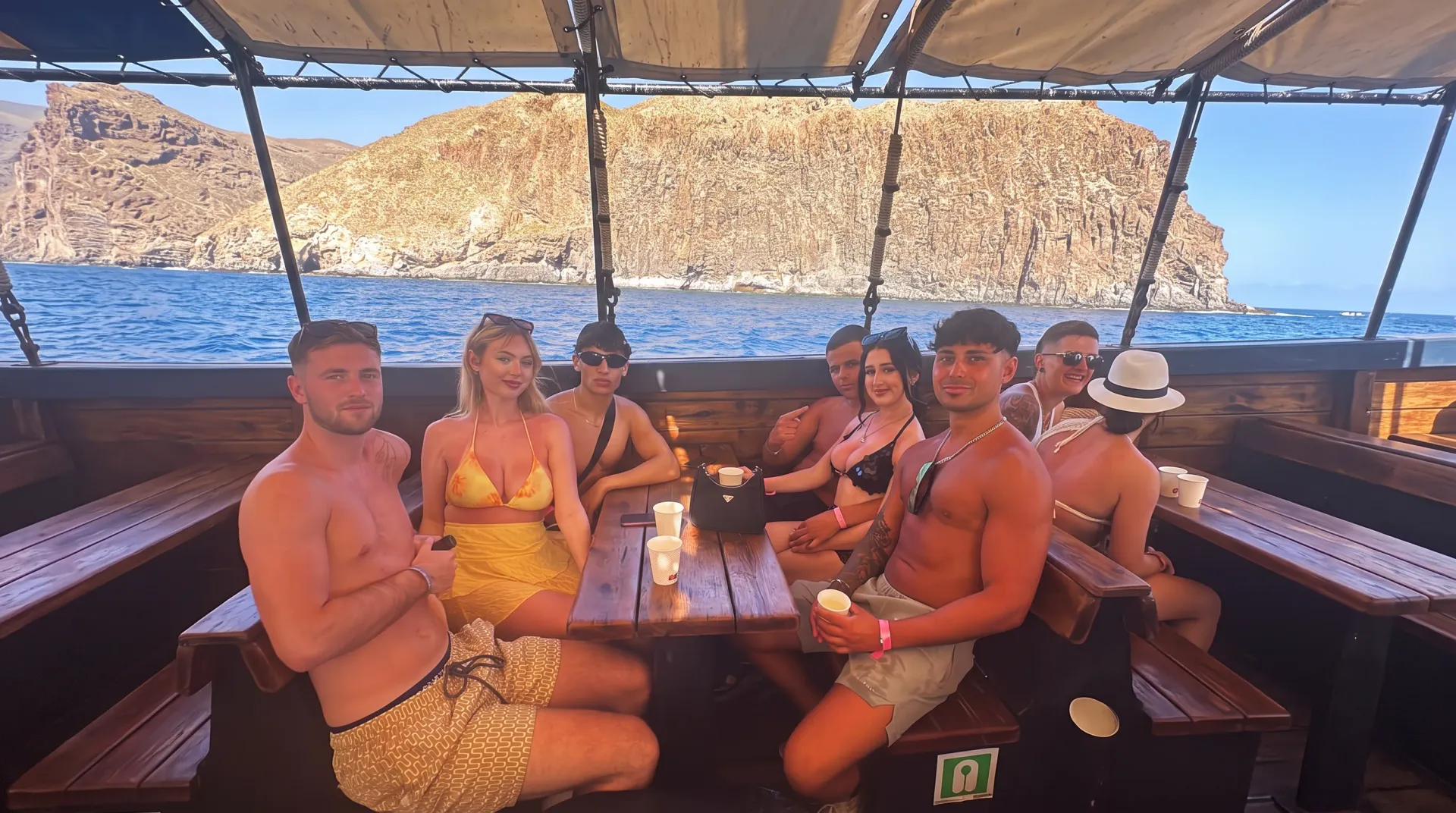 Group of guests relaxing on wooden benches aboard boat near rocky cliffs