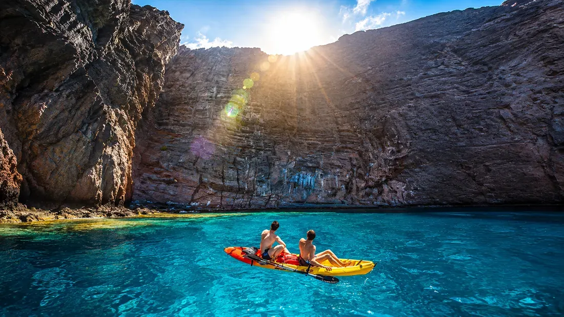 Kayakers paddling in turquoise water near rocky cliffs at sunset