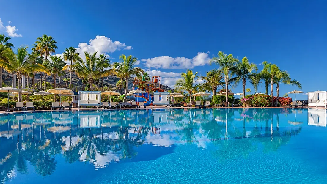 Luxury resort pool with palm trees, umbrellas, and blue sky reflections