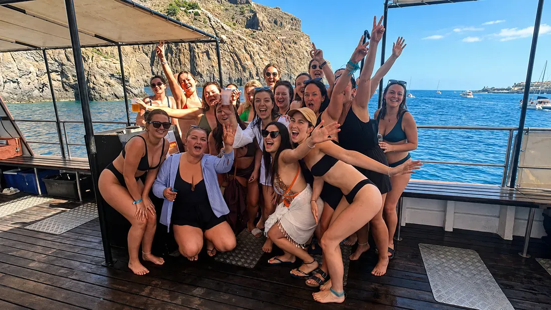 Group of women celebrating on boat deck with ocean and cliffs in background